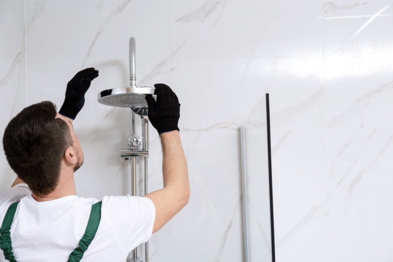 A plumber wearing gloves as he inspects a shower head in a bathroom with while marble walls.