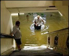 Two people walking in a flooded room at the bottom of a flight of stairs.