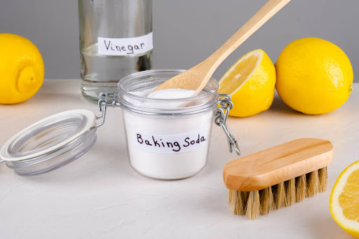 A container of baking soda and a glass of vinegar on a table with lemons and a brush.